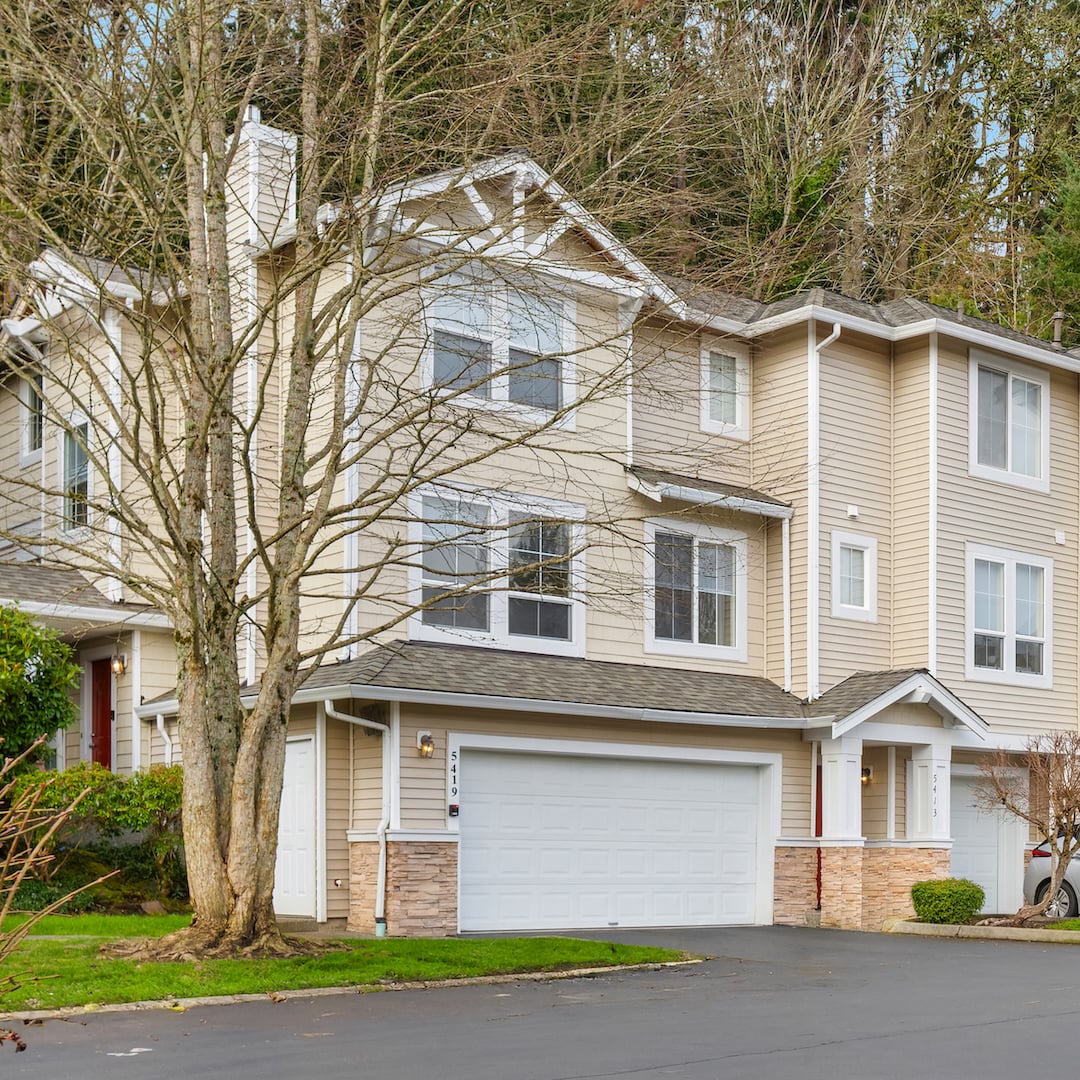Exterior view of a three-story townhome with beige siding, white garage door, stone accents, and mature trees backdrop.
