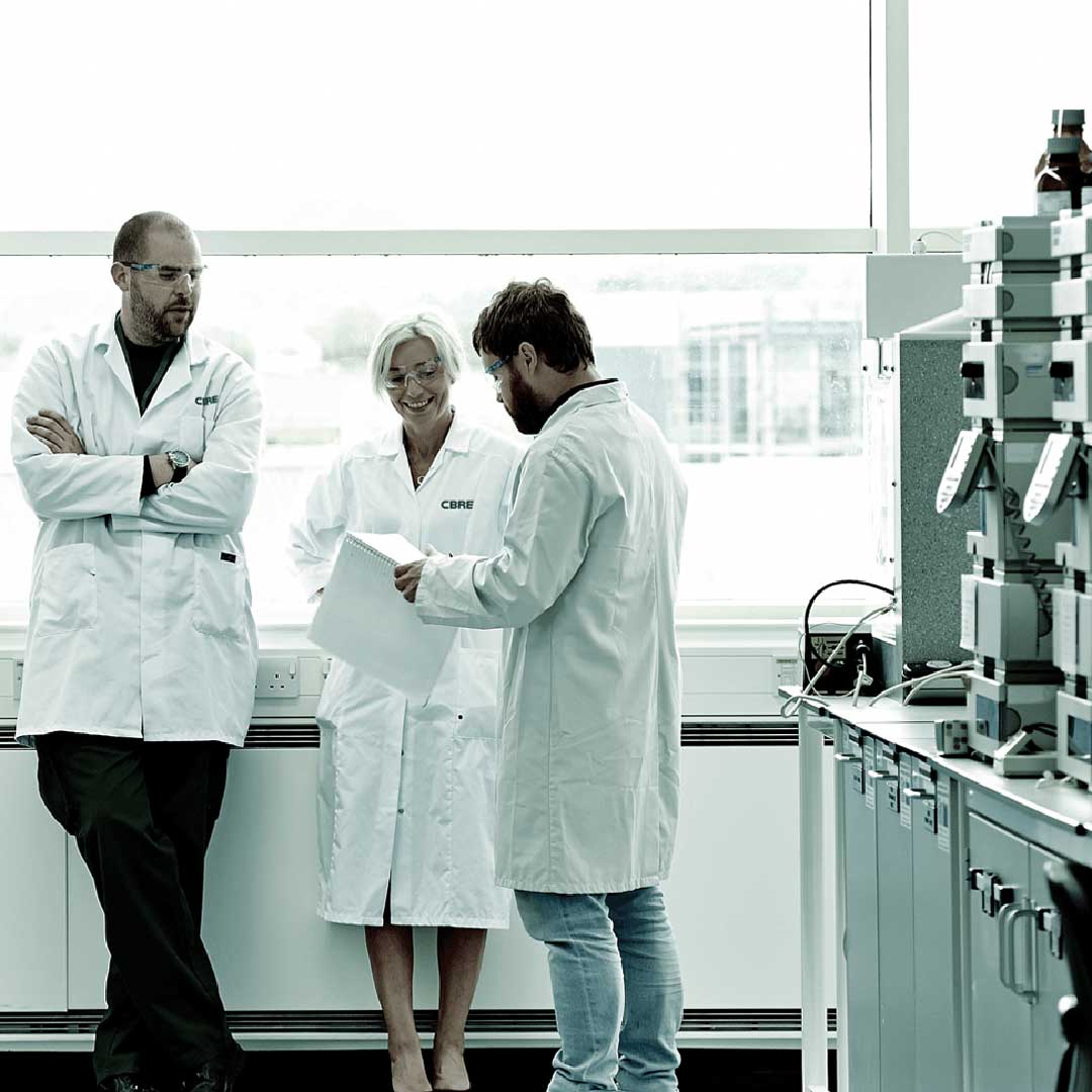 Three scientists in CBRE-branded lab coats reviewing documents in a laboratory, representing CBRE's 2026 San Diego life sciences talent report