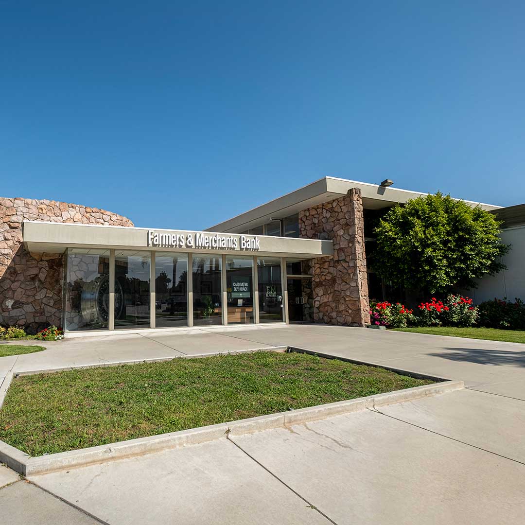 Farmers & Merchants Bank branch exterior with glass entrance, stone facade, landscaped frontage and clear blue sky.