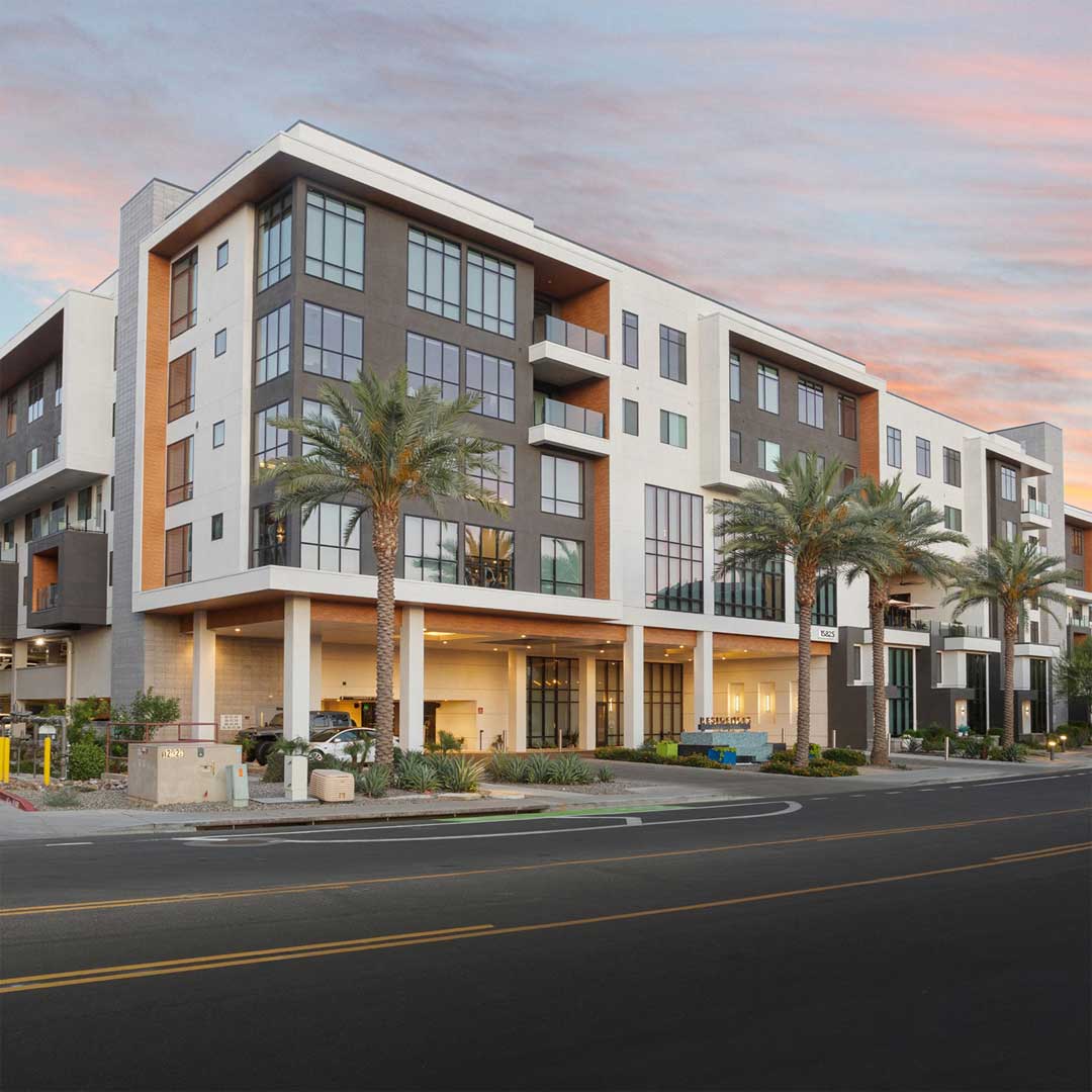 Modern multifamily apartment building in Scottsdale AZ with balconies, street frontage, palm trees, and covered parking entrance at sunset.