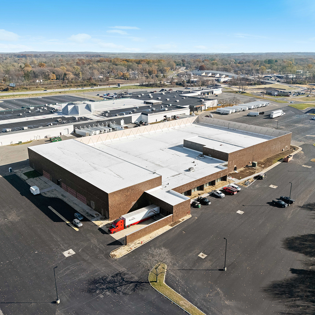 Aerial view of a large brick industrial warehouse and distribution center with a semi-truck at the loading dock, expansive surface parking, and a suburban commercial corridor with fall foliage in the background