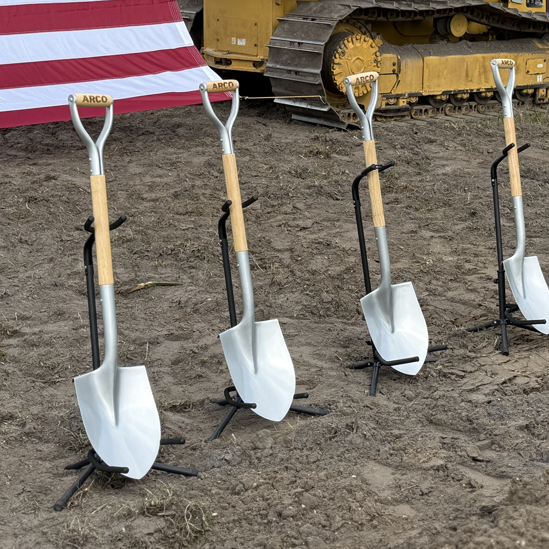 Four ARCO ceremonial groundbreaking shovels displayed on stands in bare dirt with an American flag and CAT bulldozer in the background.
