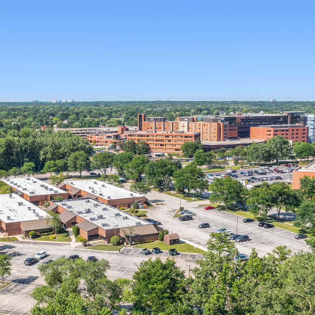 Aerial view of a large commercial campus with multiple brick buildings and surface parking lots in a suburban setting in Elk Grove Village, Illinois.