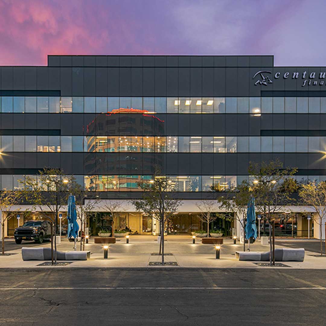 Exterior view of a modern office building with glass façade, landscaped courtyard, and illuminated interiors at dusk in Anaheim, California.