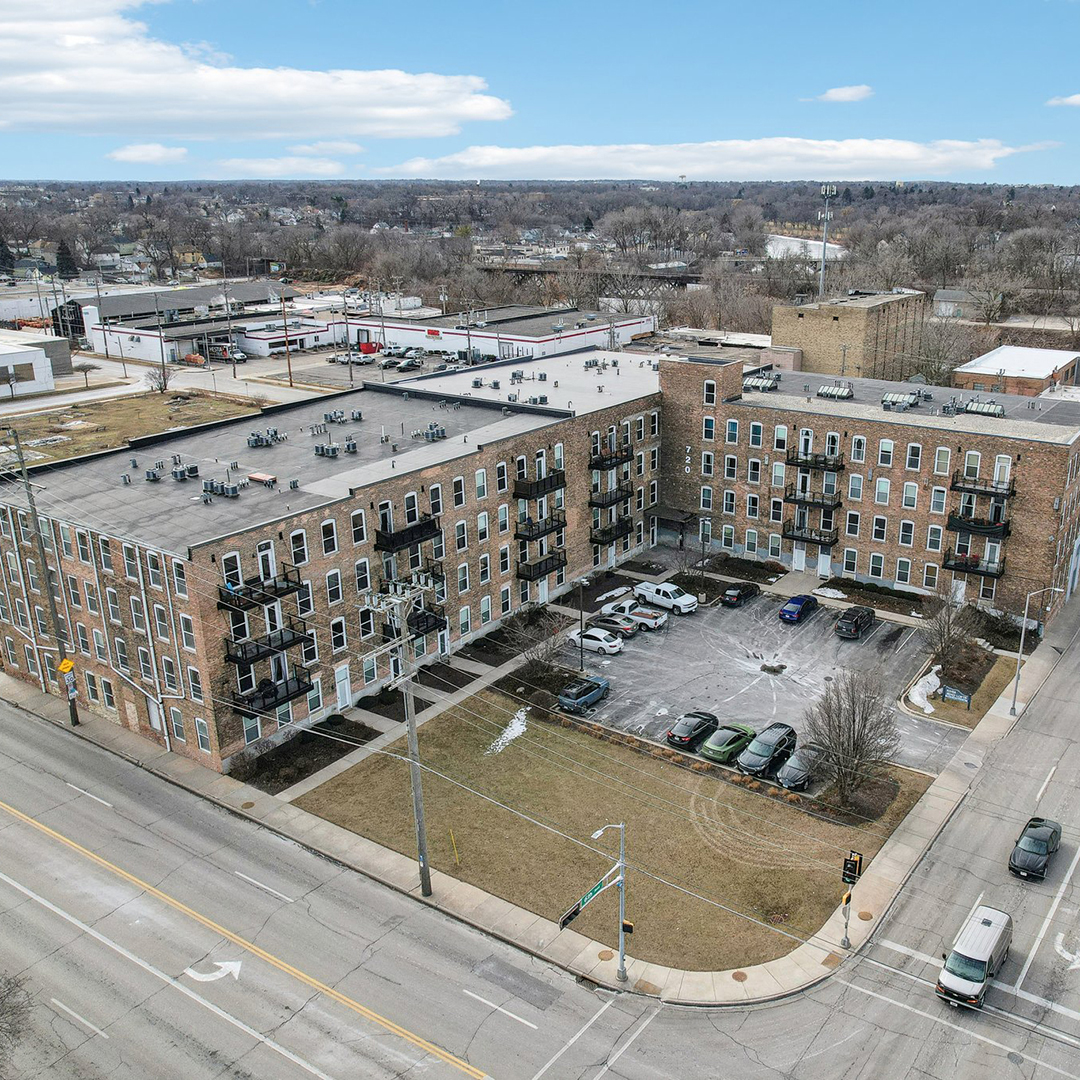 Aerial view of a mid‑rise brick apartment building with balconies and a surface parking lot at a street corner.
