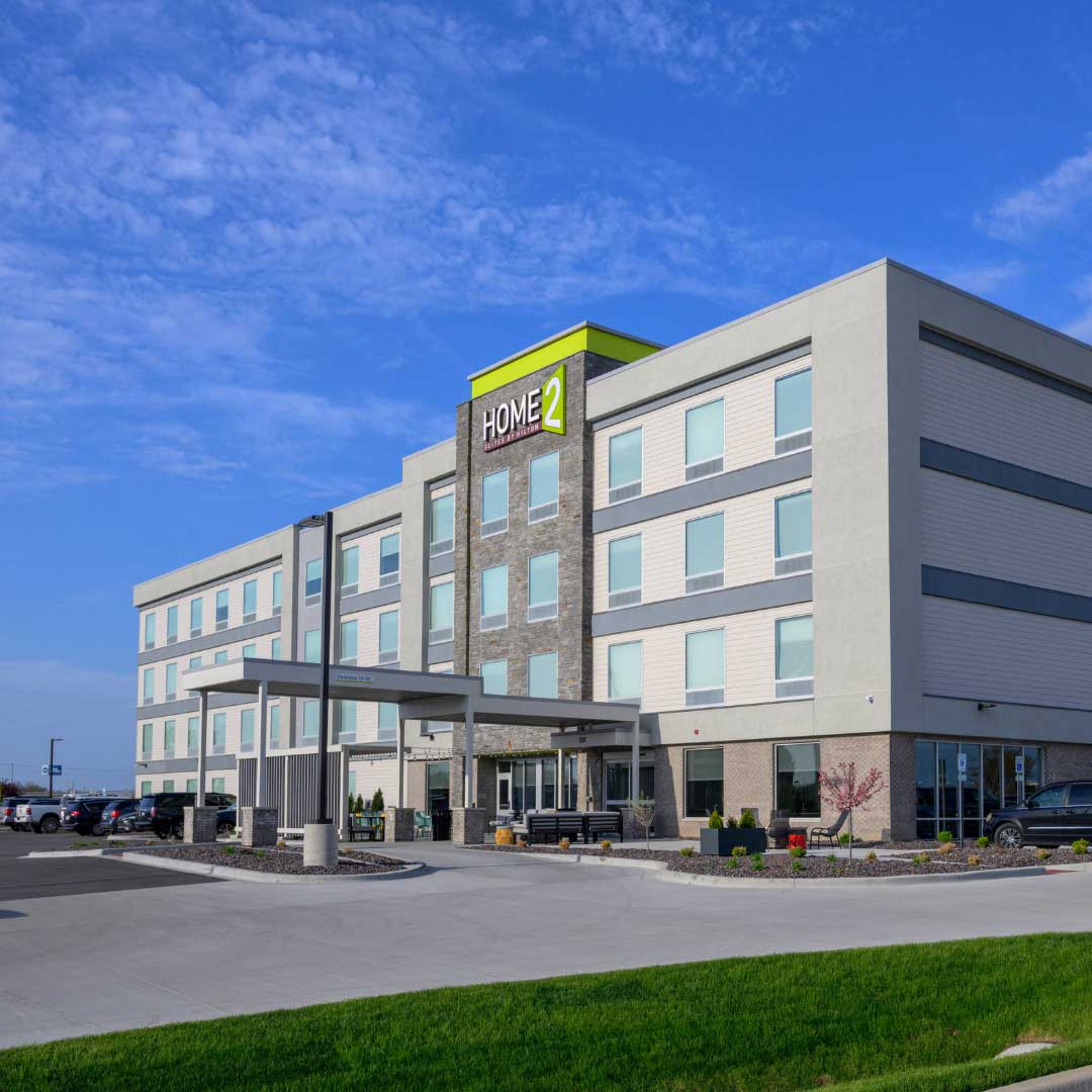 Exterior view of Home2 Suites by Hilton hotel featuring modern design, main entrance, and landscaped surroundings under a clear blue sky.