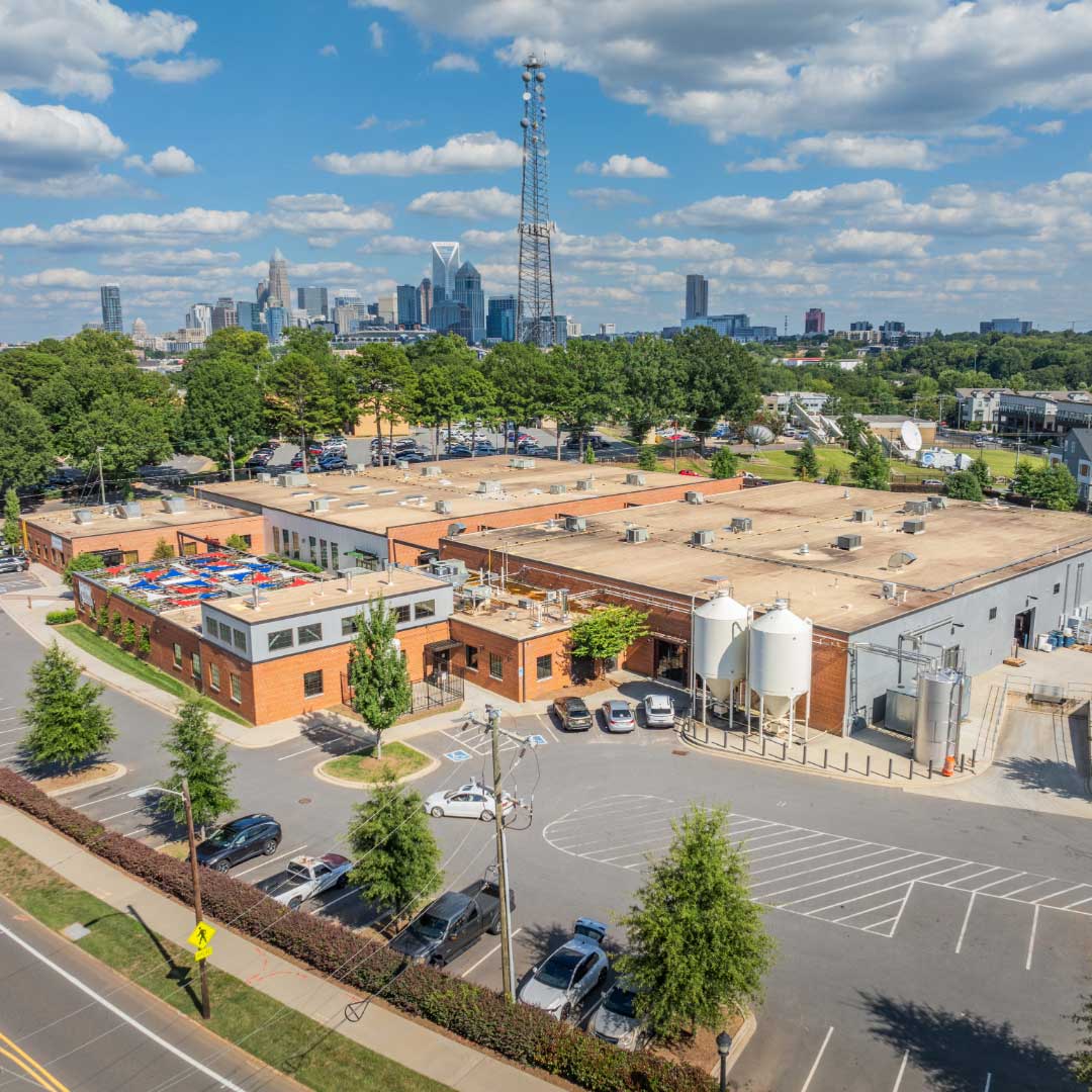 Aerial view of an industrial property with brick buildings, parking areas, and city skyline in the background under a clear sky.
