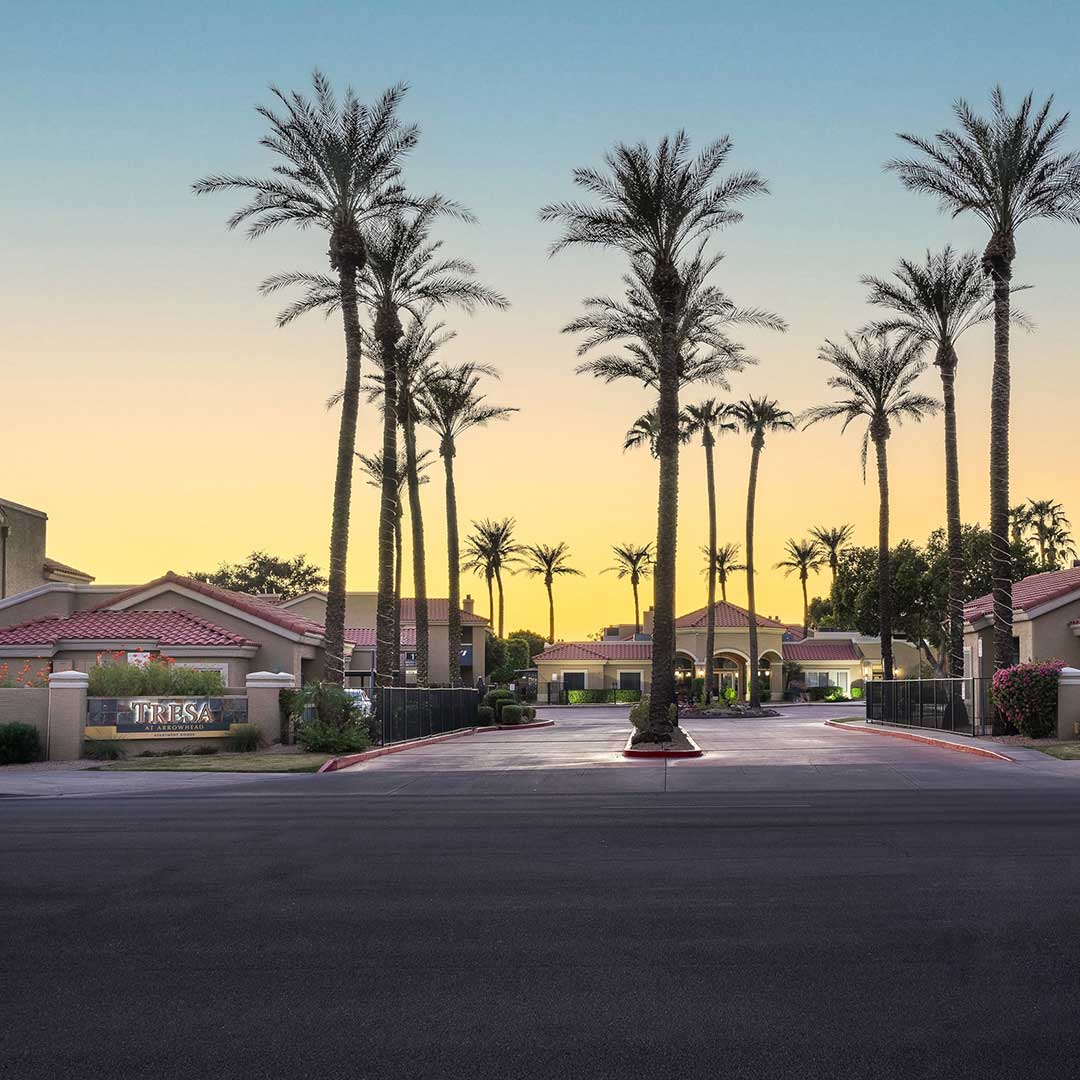 Palm-lined entrance to a multifamily residential community with tiled-roof buildings and a central driveway at sunset in Glendale, Arizona.