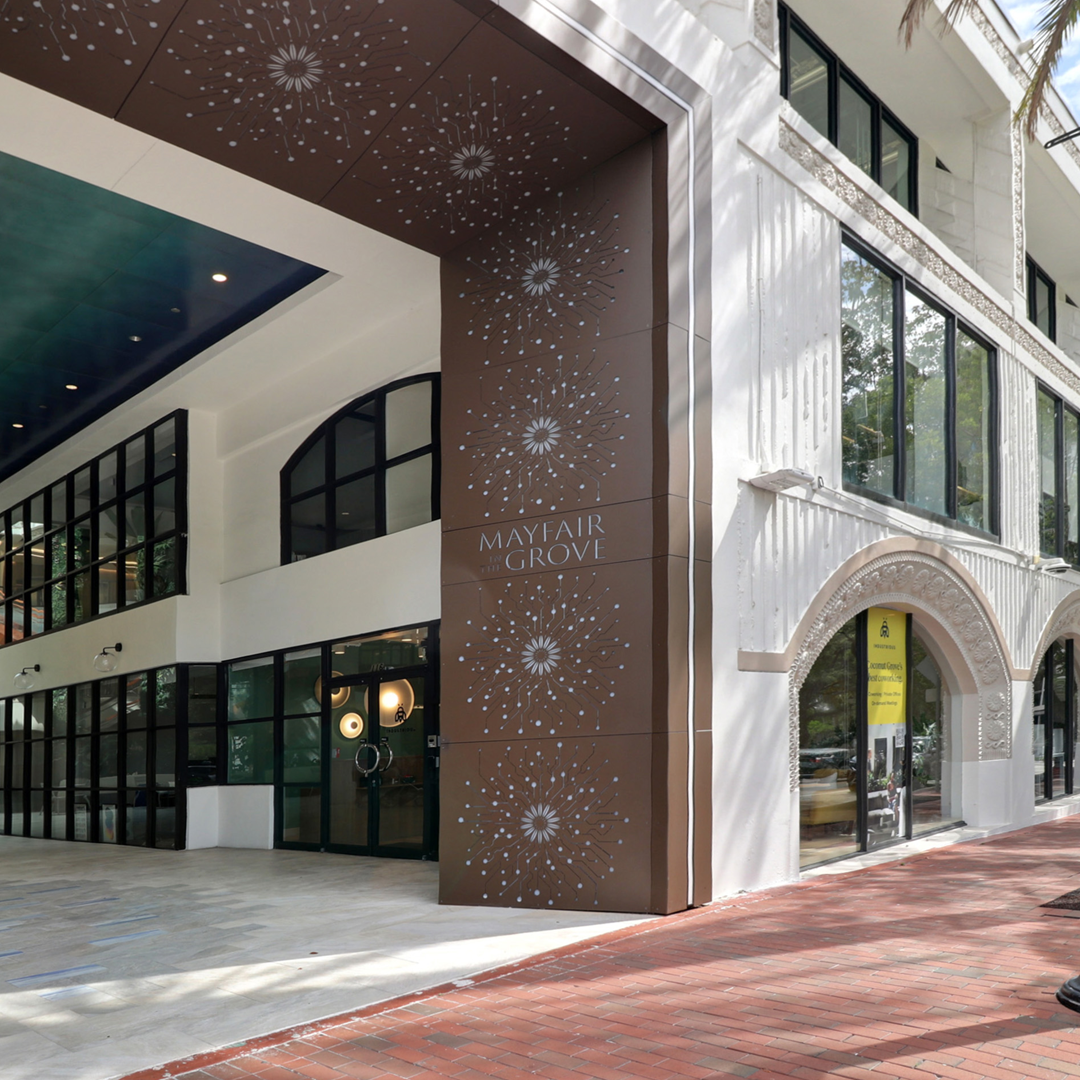Street-level exterior of the Mayfair Grove mixed-use building with retail storefronts, decorative façade, and pedestrian walkway.