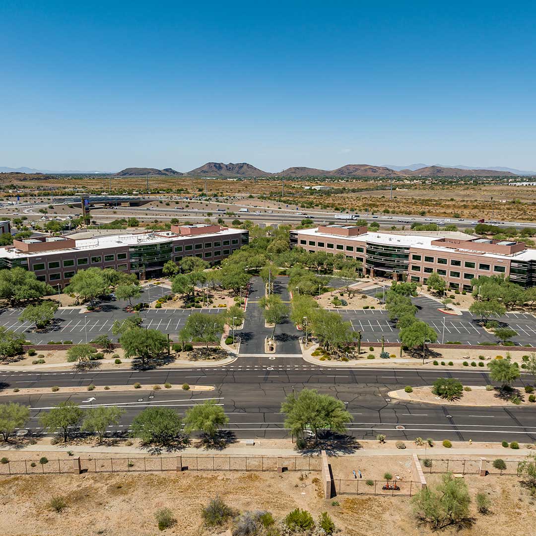 Aerial view of a multi-building office campus with landscaped parking areas, wide roads, and desert mountains in the background.