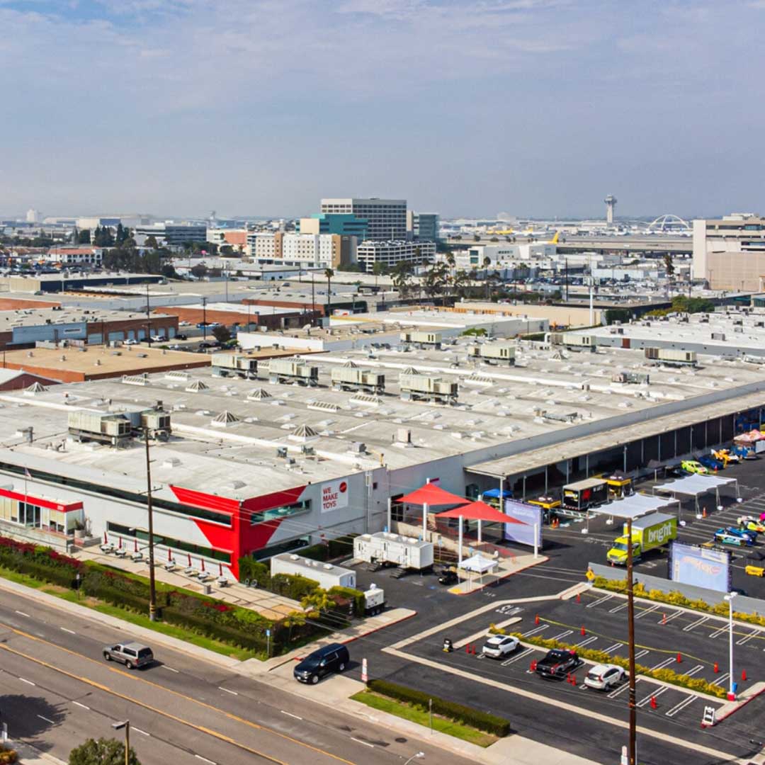 Aerial view of a large industrial warehouse complex with parking areas, trucks, and surrounding commercial buildings in an urban setting.
