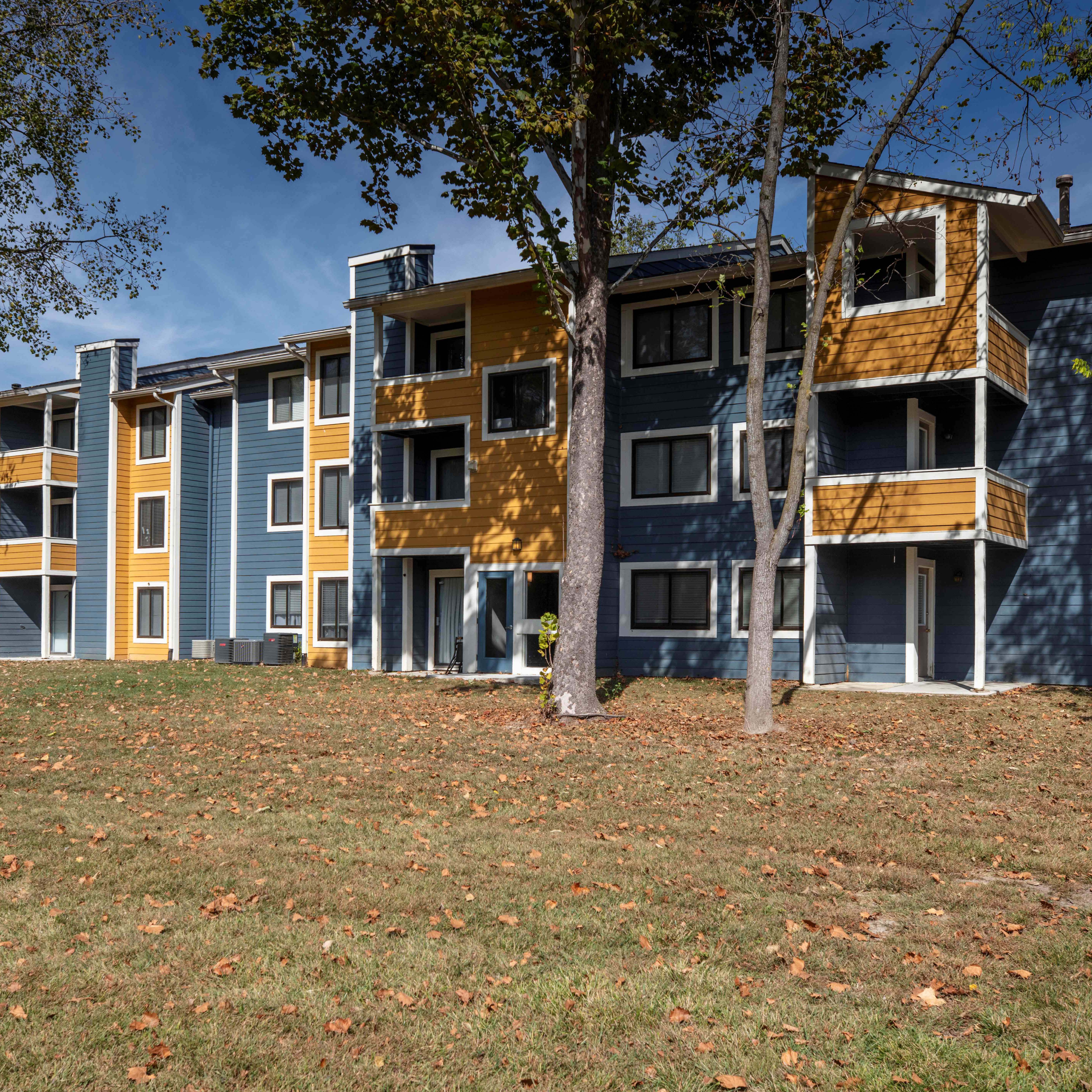 Exterior view of the Deercross multifamily apartment building with balconies, blue and yellow siding, trees, and landscaped lawn.