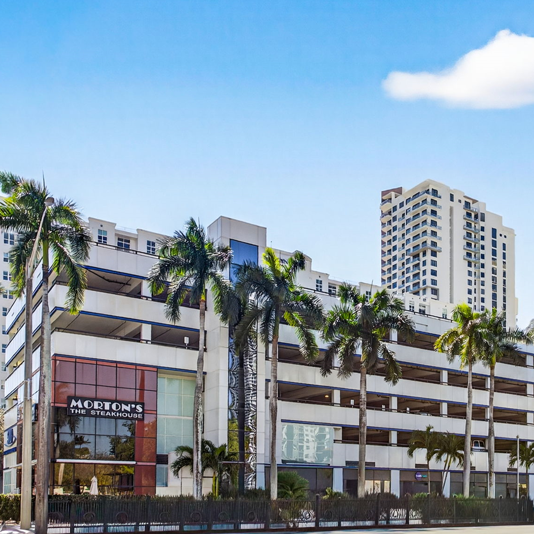Exterior view of a mixed-use commercial building with a multi-level parking structure, palm trees, and Morton’s The Steakhouse at street level.