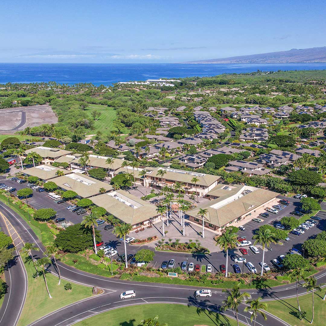 Aerial view of a coastal commercial complex surrounded by greenery, nearby residential areas, and ocean in the background.