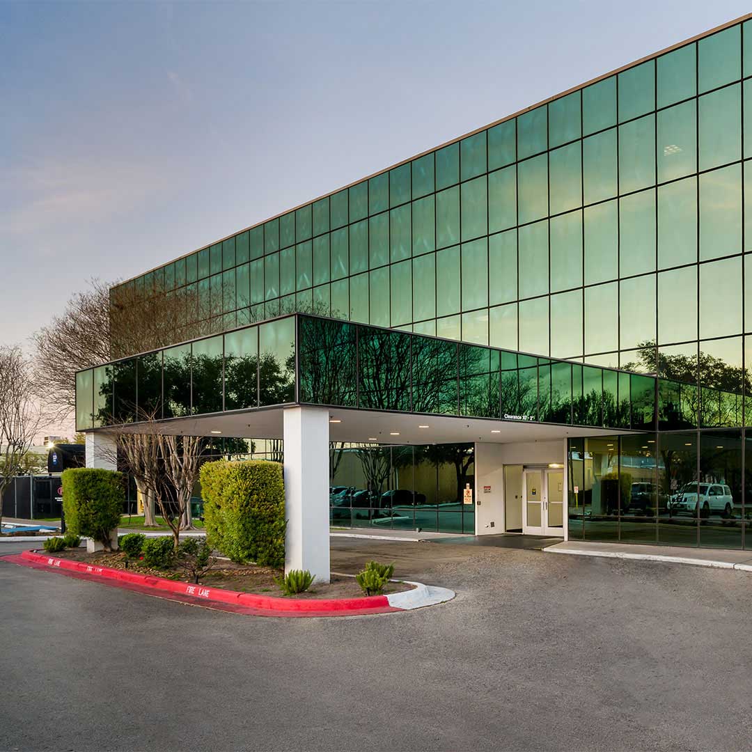 Modern glass office building exterior with reflective green windows, covered entrance, landscaped driveway, and contemporary commercial architecture.