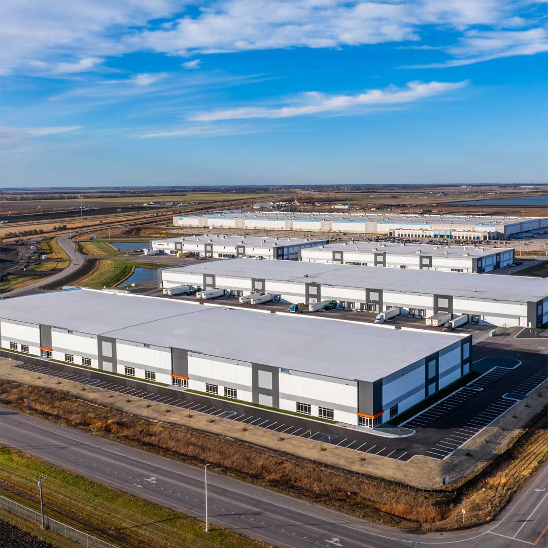Modern industrial warehouse complex with loading docks, trucks, and large logistics distribution center in a rural commercial area under blue sky.