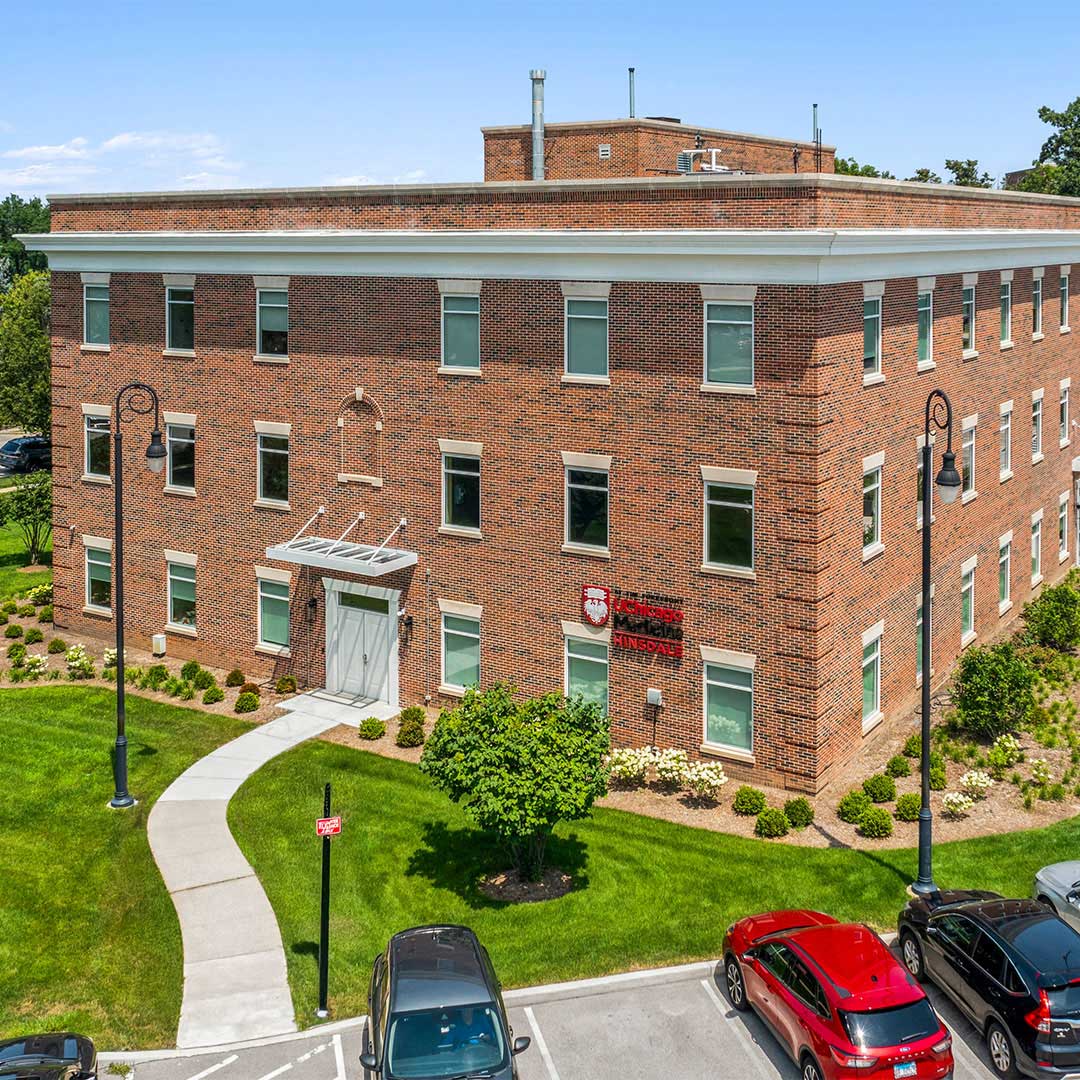 Three-story red brick building with multiple windows, landscaped lawn, walkway, parked cars, and a clear blue sky.