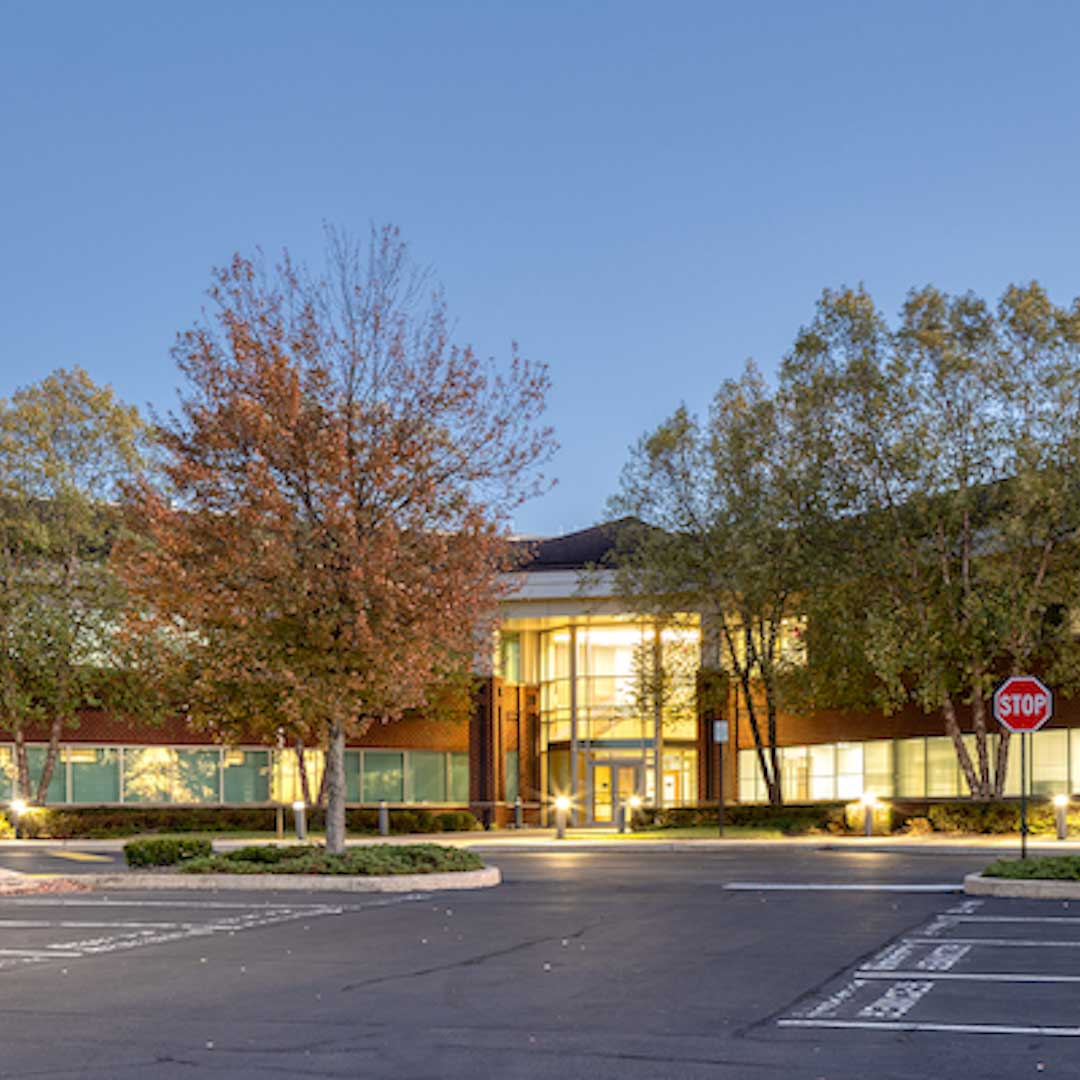 Modern office building exterior with illuminated entrance, autumn trees, and empty parking lot at dusk under a clear blue sky.