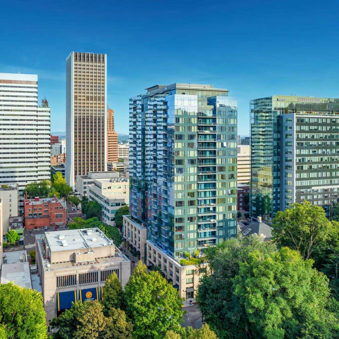 Modern high-rise residential and office buildings with glass facades in a vibrant urban cityscape, surrounded by trees and streets under a clear blue sky.