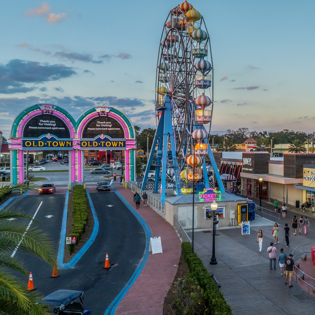 Aerial dusk view of Old Town Kissimmee featuring a colorful Ferris wheel, illuminated entrance arches, and visitors walking along the promenade.