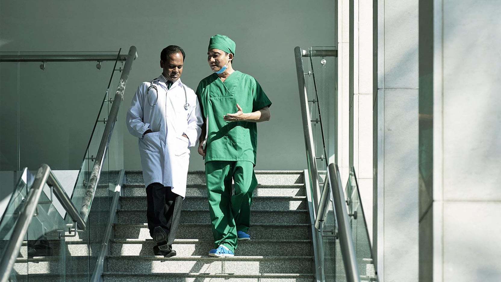 Two medical professionals, a doctor in a lab coat and a surgeon in green scrubs, walk down a modern hospital staircase, discussing healthcare.