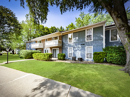 Exterior of Canterbury Green garden-style apartment community featuring two-story brick and blue siding buildings with mature trees and manicured lawn.