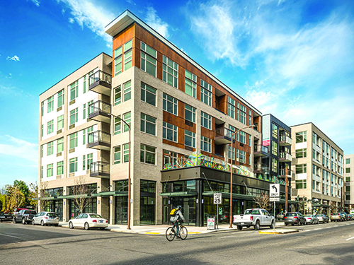 Street corner exterior of Peloton urban mixed-use apartment building with rust-panel and brick facade, ground-floor retail, and bike-friendly streetscape.
