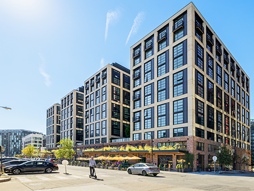 Street-level exterior of The Batley urban mixed-use apartment building featuring dark metal and masonry facade with ground-floor restaurant patios.