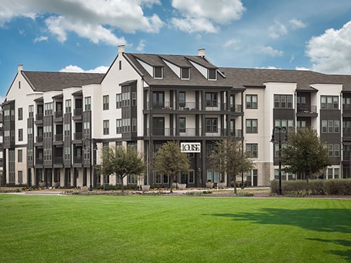 Exterior of The Louise apartment community showing a four-story dark and white facade with balconies, ground-floor signage, and manicured green lawn.