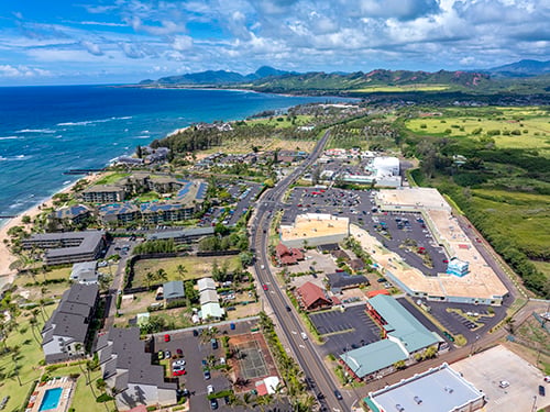 Aerial view of a coastal shopping center featured in the Epic Retail Portfolio, showing retail buildings, parking lots, a main highway, and the ocean coastline with mountains in the distance.