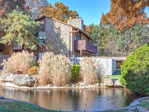 Kenilworth property featuring a multi-story stone and wood residential building with a natural pond, ornamental grasses, and autumn foliage