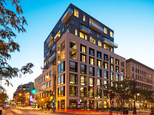 Sugar Block mixed-use residential and retail building at a downtown street corner illuminated at dusk with warm window lighting and city traffic