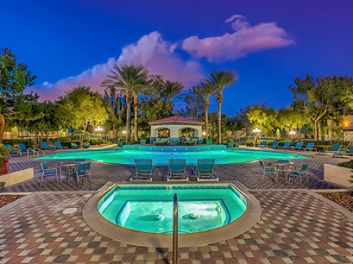 Resort-style outdoor swimming pool and spa at an apartment community, featuring palm trees, lounge seating, and evening lighting.