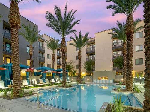 Resort-style swimming pool courtyard at a modern apartment community with palm trees, lounge seating, and mid-rise residential buildings at sunset.