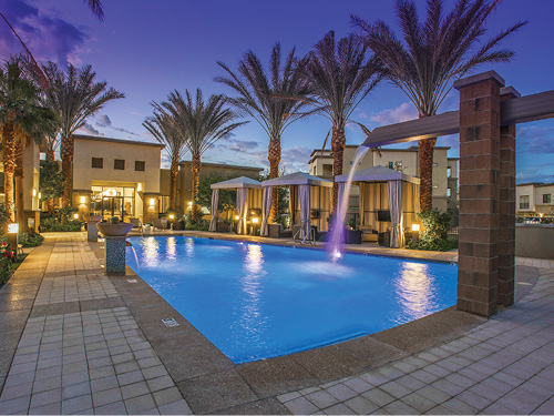 Twilight photo of Cadia Crossing apartment community resort-style pool with water fountain feature, palm trees, private cabanas, and warm accent lighting.
