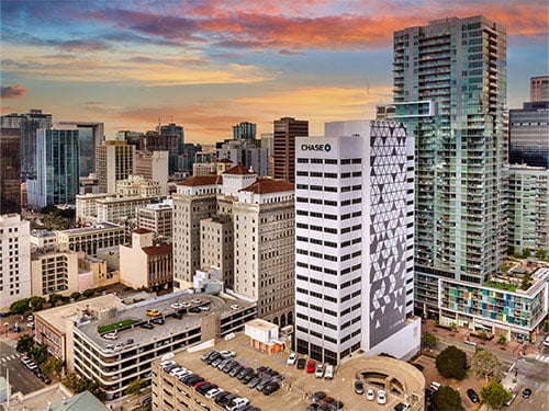 A white high-rise office building with geometric exterior design set within a dense downtown skyline at sunset.
