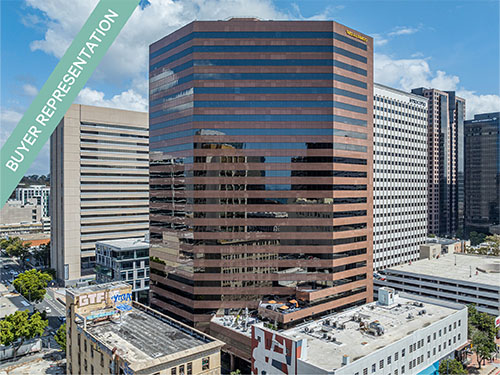 A reflective glass office skyscraper in an urban core, captured from above with surrounding mid-rise buildings.
